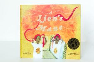 Cover of A Lion's Mane. A young boy stands in front of a mirror to tie a red dastaar (Sikh turban) and sees whiskers on his face in the mirror. Written by Navjot Kaur and Illustrated by Jaspreet Sandhu. ©Saffron Press